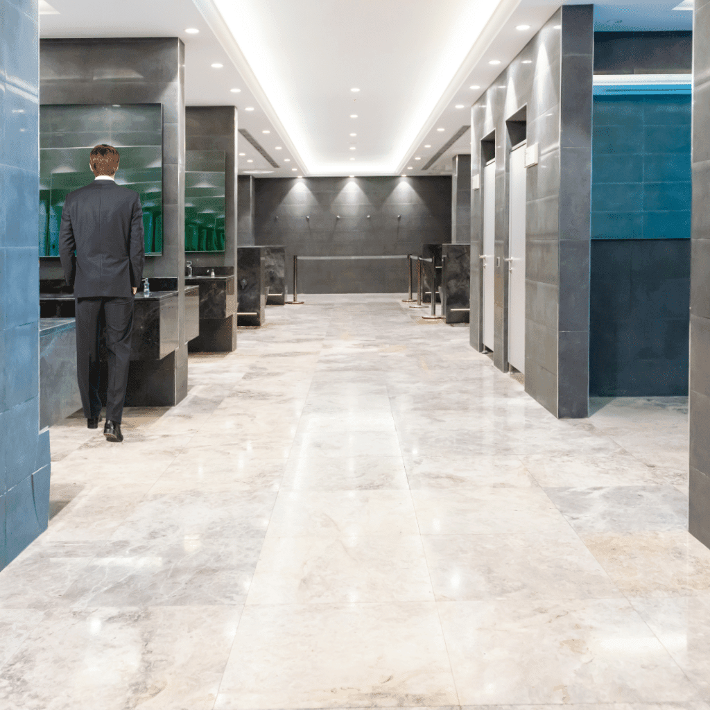 A businessman in a dark suit walking away from the viewer down a modern, upscale public restroom with polished marble floors, dark gray tile walls, recessed lighting, and teal-colored accents. The space features sleek fixtures and a long corridor perspective.